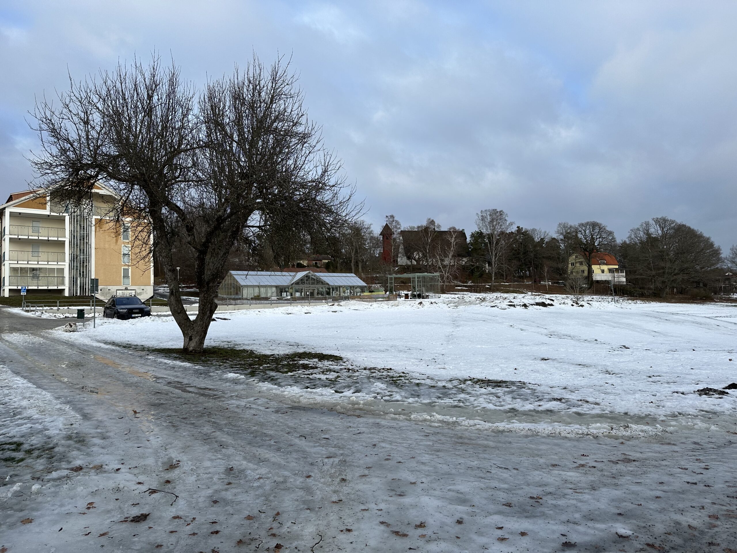 Isig gångväg framför snöklädd gräsmatta. I bakgrunden hus, växthus och Sköndals kyrka. 