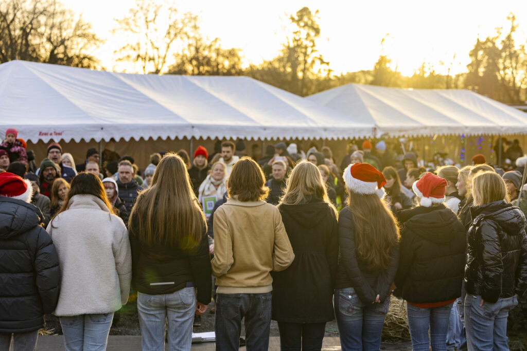Ryggarna på en grupp ungdomar som uppträder med julsånger framför åhörare på Stora Sköndals julmarknad. I bakgrunden ses vita utställartält.