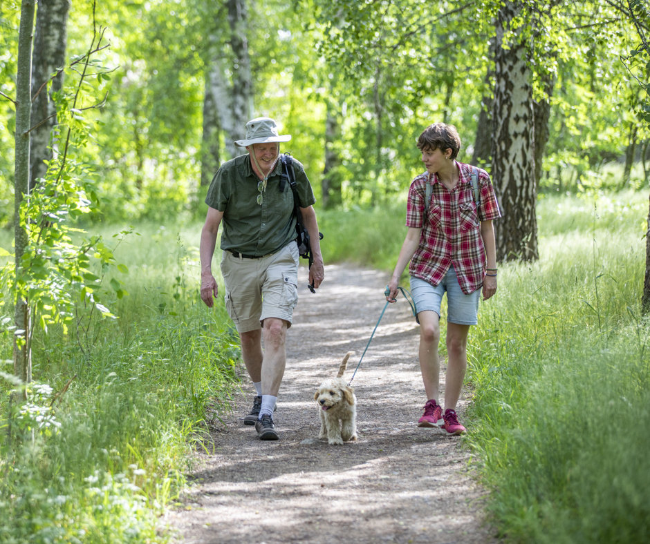 En man och en ung kvinna promenerar med en hund på en grusväg, omgivna av lummig lövskog.