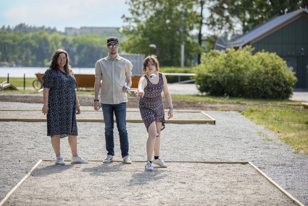 Tre personer spelar boule en solig sommardag. I bakgrunden grönska och en sjö.