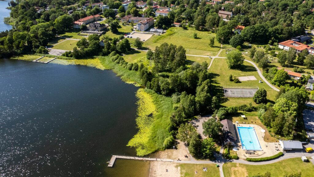 Vy över strandområde med badbryggor och sandstrand, bassäng, banor för tennis och volleyboll, solbrygga och gröna gräsytor. 
