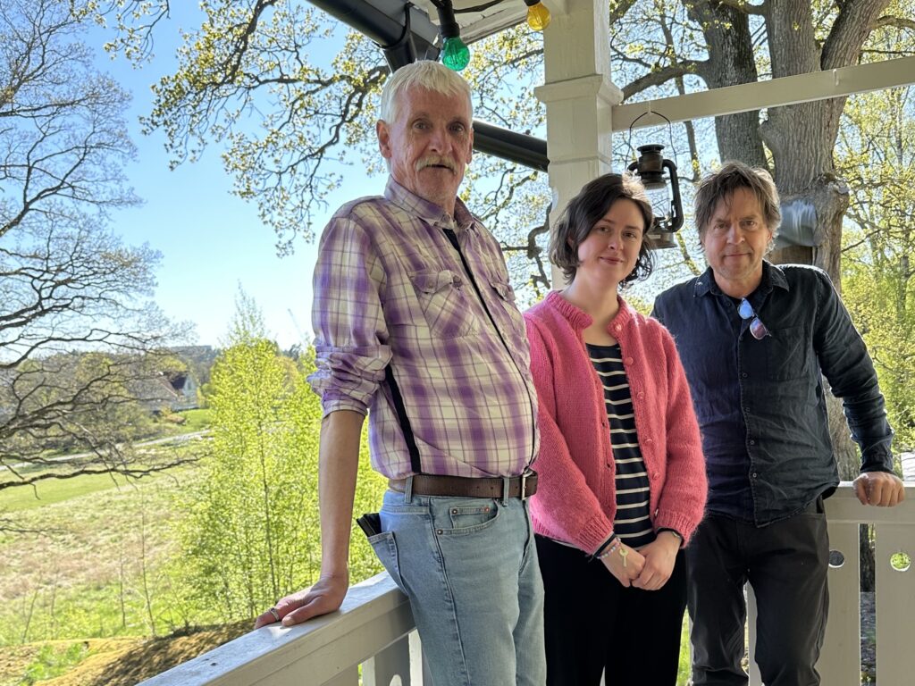 Tre personer på veranda, med grönskande bakgrund.